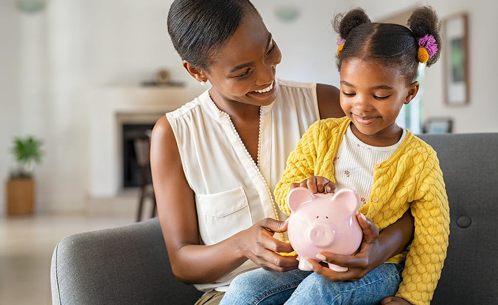 A mother and daughter sit together and smile. They’re looking at a piggy bank.